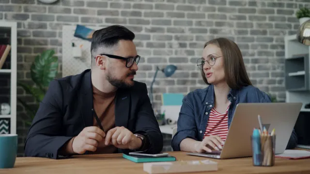 Dvaja kolegovia sa rozprávajú pri stole s notebookom v kancelárii