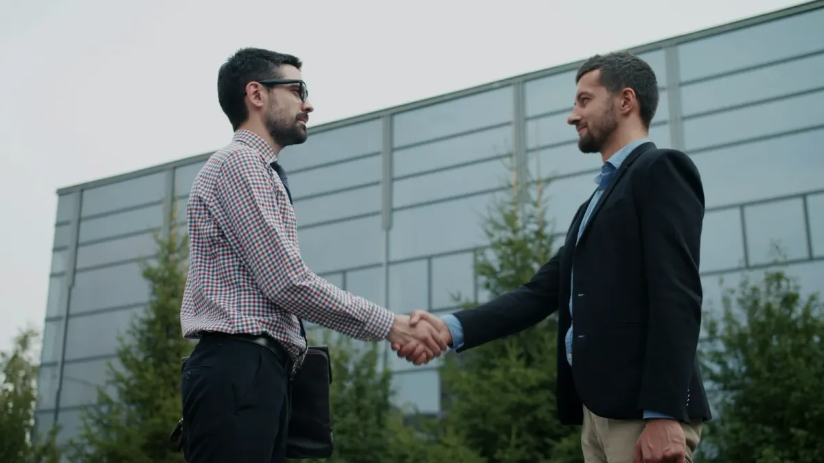 Dvaja muži v obleku si podávajú ruky pred modernou administratívnou budovou