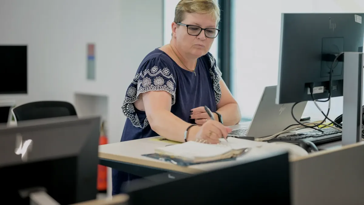 Žena sústredene píše poznámky do zošita pri pracovnom stole s monitormi