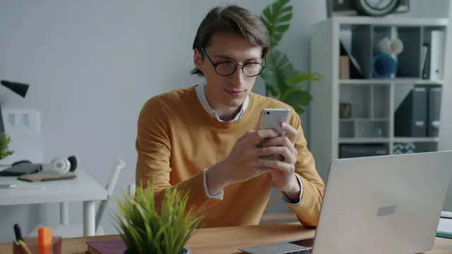 Muž pri stole s notebookom sa sústredene pozerá do telefónu